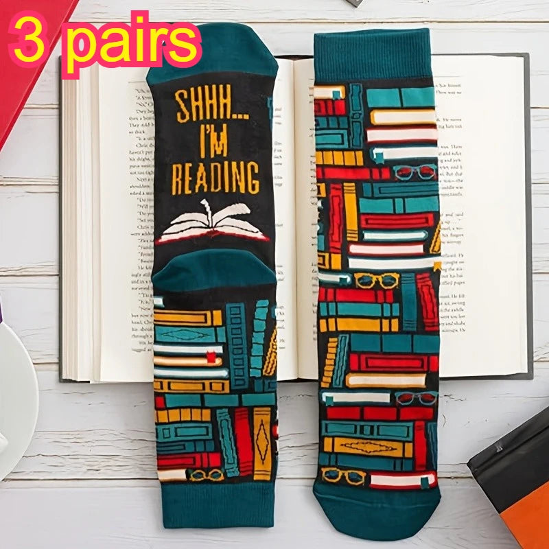 Three pairs of novelty socks displayed on an open book. One sock has a dark teal and black background with yellow text reading 'SHHH.