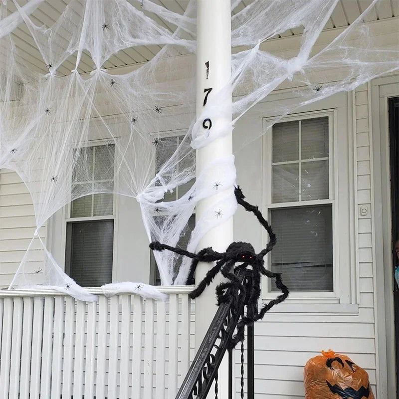 Halloween porch decorations featuring white fake cobwebbing draped across a house exterior, a large black fuzzy spider on a white porch post, and an orange jack-o'-lantern bag on the ground.