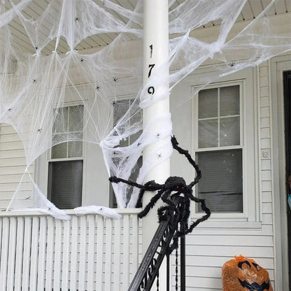 Halloween porch decorations featuring white fake cobwebbing draped across a house exterior, a large black fuzzy spider on a white porch post, and an orange jack-o'-lantern bag on the ground.