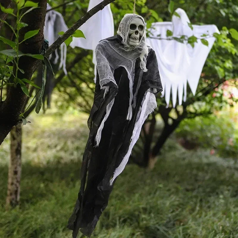 A hanging Halloween decoration shaped like a skeletal grim reaper in a tattered black and gray robe, photographed outdoors among trees and grass. White ghost-like decorations are also visible in the background.