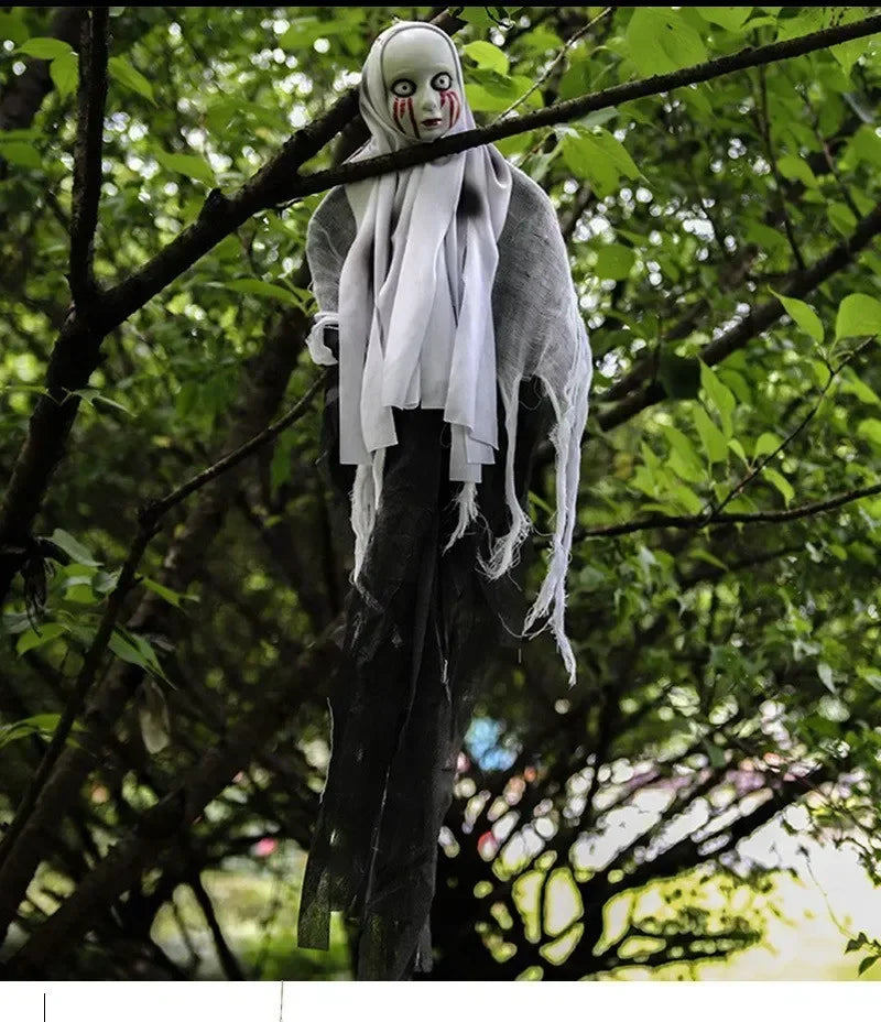 A hanging Halloween decoration shaped like a ghostly figure with a white mask, black body, and tattered white cloth, suspended from a tree branch in a real outdoor setting with green leaves.