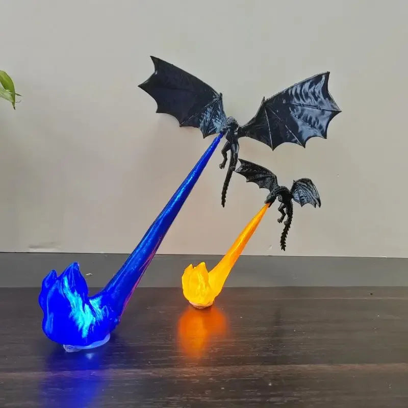 Two black dragon figurines with spread wings, each mounted on a flame-shaped base that glows blue or orange, displayed on a dark wooden surface against a plain beige wall.