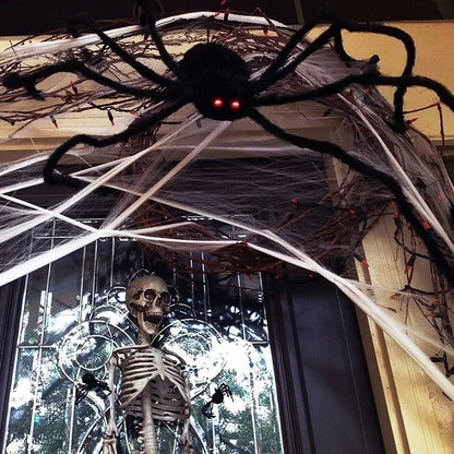 Halloween decoration set featuring a large black fuzzy spider with glowing red eyes draped over a web structure, with a skeleton prop standing below in front of a window.