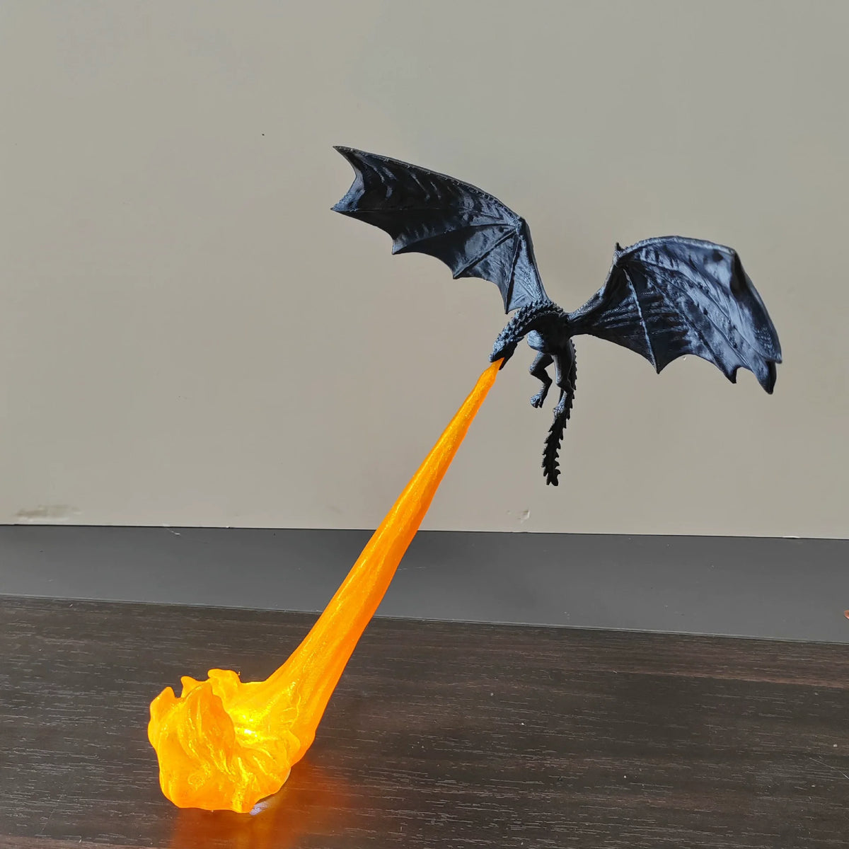 A black dragon figurine with spread wings, breathing a bright orange stream of fire that ends in a glowing flame on a dark tabletop against a plain beige wall.