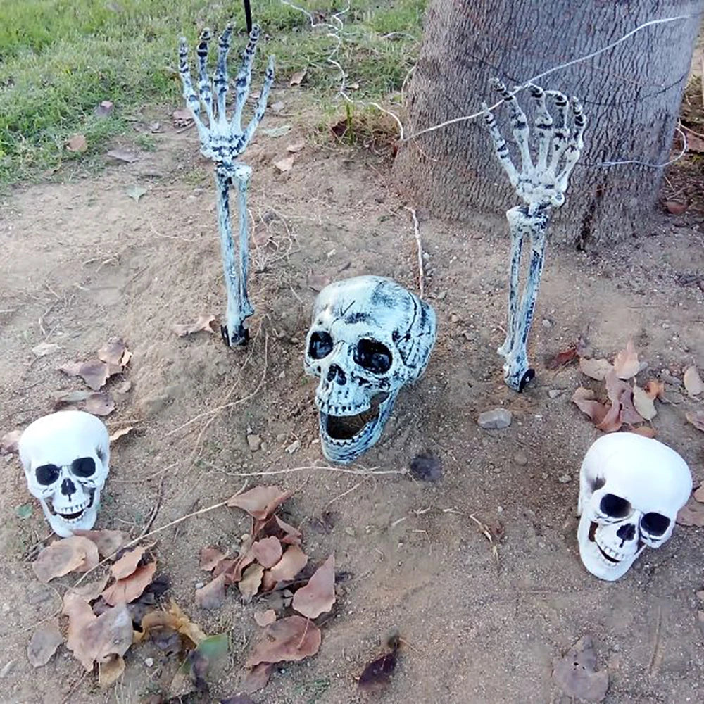 Three Halloween skeleton decorations displayed outdoors on dirt ground near a tree trunk, including two upright skeletal hands and a central skull with two smaller skull props at the lower left and right.