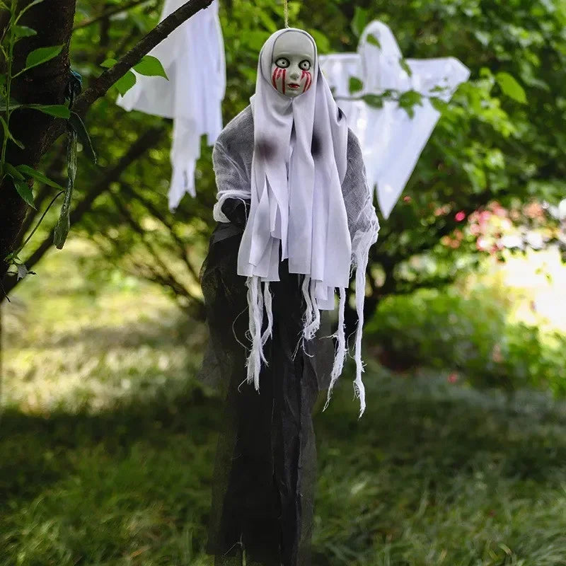 A Halloween hanging ghost decoration stands outdoors in a grassy yard. The ghost figure has a pale gray face with red painted tears, a tattered white sheet-like head covering, a dark gray draped body, and a long black hanging skirt.